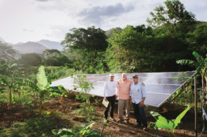 Three solar energy professionals stand smiling in front of a newly installed solar panel in rural Colombia