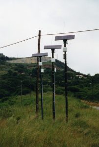 Three solar panels sit on top of poles in a mountainous landscape