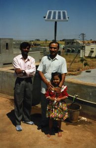 Two men and a child smile in front of a new solar panel in rural India