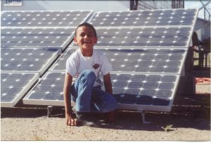 A boy sits in front on a solar panel smiling on Navajo Nation land
