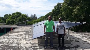 Two men stand in front of newly installed solar panels from the Solar Electric Light Fund (SELF) in Haiti