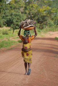 Woman with a child strapped to her back walk down a dirt road carrying wood on her head